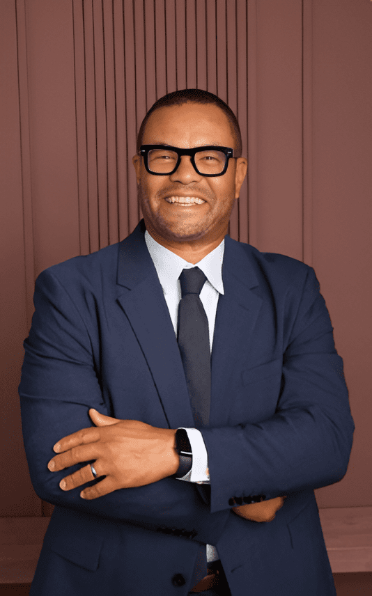 Confident man in a navy suit and glasses smiling with arms crossed.